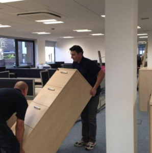 Two commercial movers dragging a shelf across the office floor. 