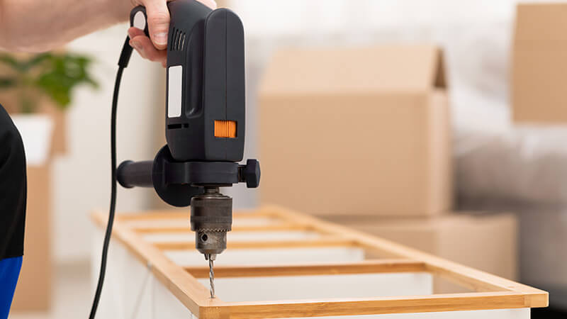 A person holding a drilling machine over a piece of furniture. 