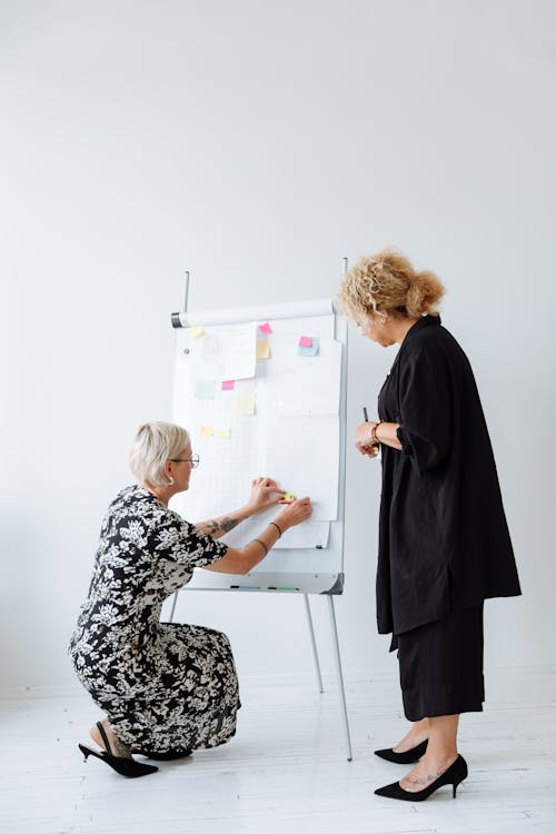 Two women collaborate using sticky notes on a whiteboard to plan an office relocation timeline.