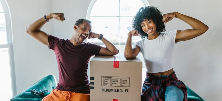 Two people sitting next to a cardboard box and smiling.